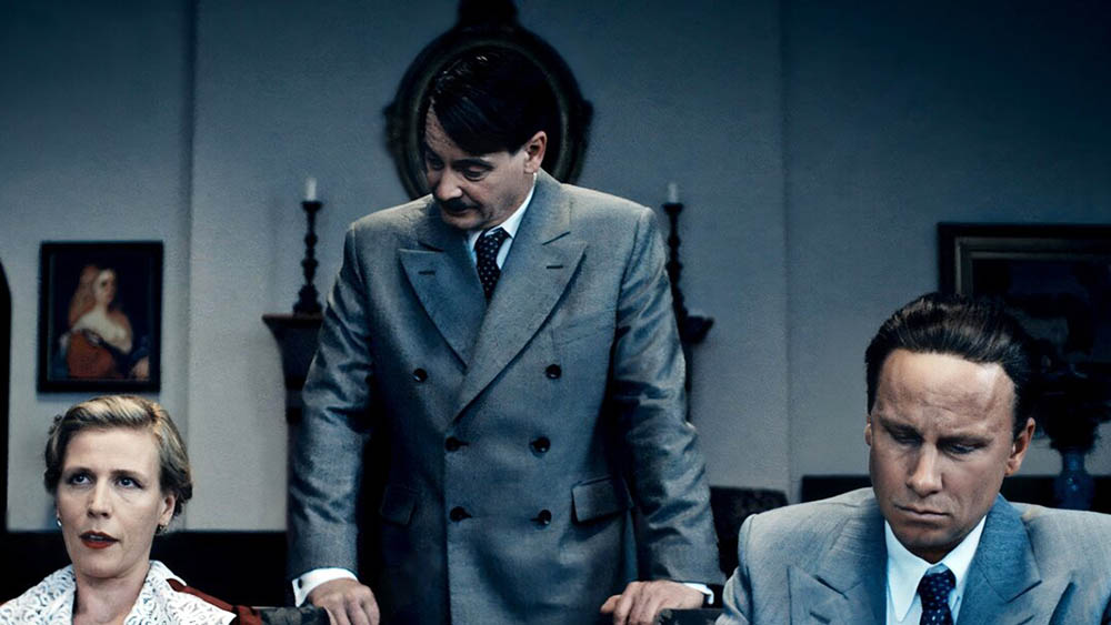 Eine Filmszene aus dem Film "Führer und Verführer". Ein Mann steht vor einem Tisch, an dem eine Frau und ein Mann sitzen.