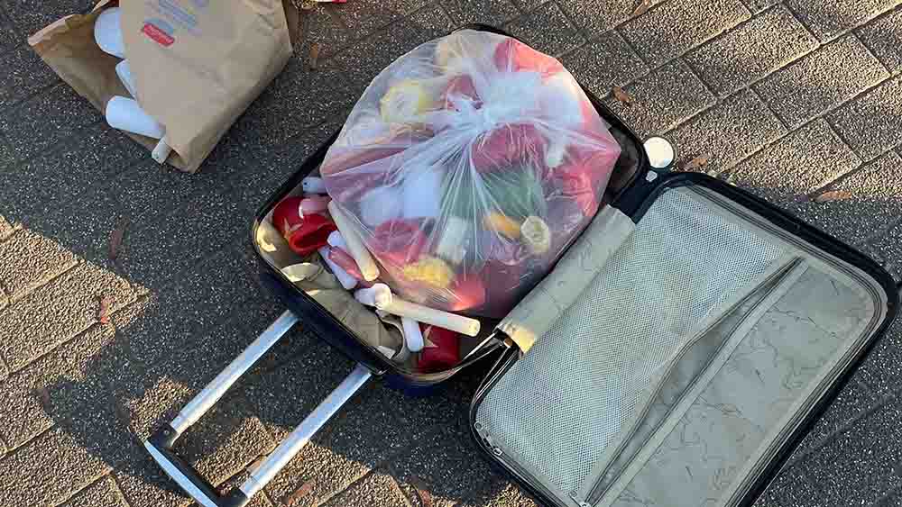 Ein Rollerkoffer liegt geöffnet da, darin sind Kerzenreste ansatzweise zu sehen. Neben dem Koffer liegt eine weiße Tüte.