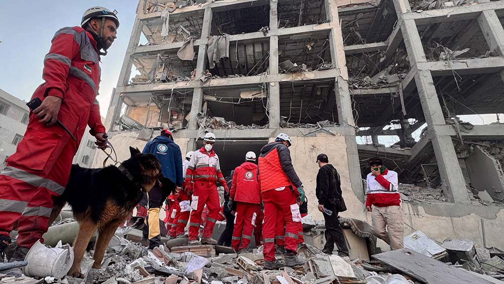 Mann in roter Kleidung mit einem Hund stehen vor einem mehrstöckigen zerstörten Haus